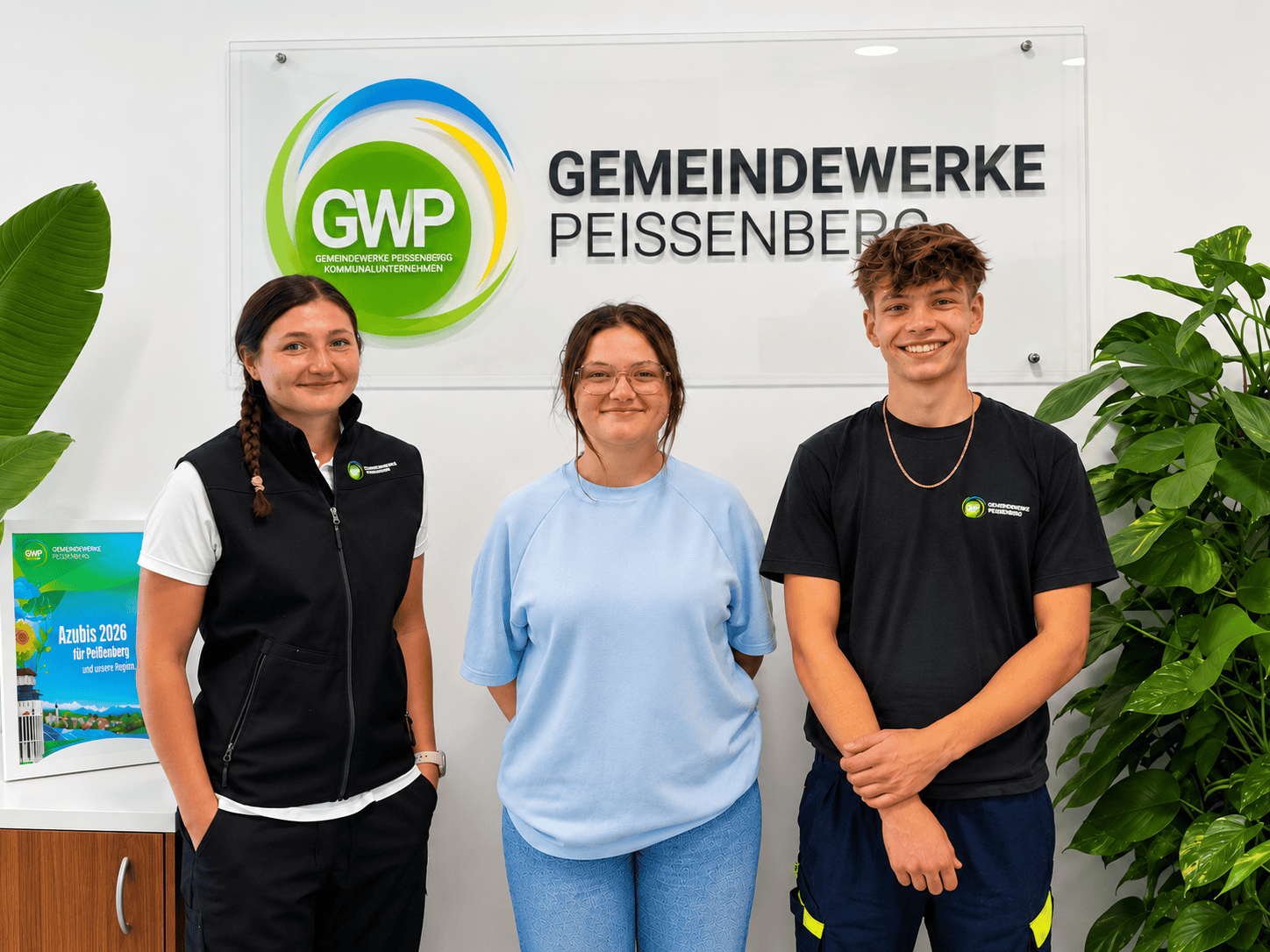 Drei Auszubildende der Gemeindewerke Peißenberg vor dem Unternehmenslogo